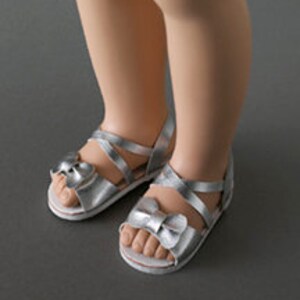 May include: Silver sandals with bow accents. The sandals have a strappy design and a white sole. The image shows the sandals on a gray surface.