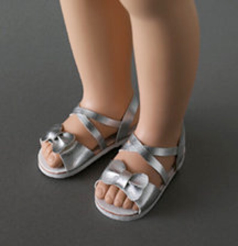 May include: Silver sandals with bow accents. The sandals have a strappy design and a white sole. The image shows the sandals on a gray surface.