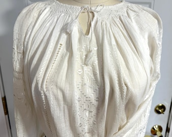 May include: Cream-colored blouse with long sleeves. The blouse features intricate embroidery and cutwork details, including a keyhole neckline with a tassel. The fabric appears to be lightweight, with vertical and geometric patterns.