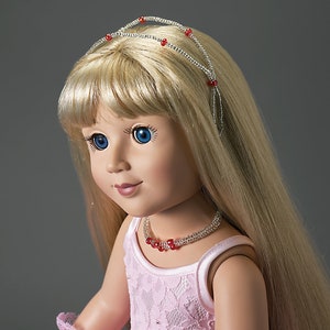 May include: A doll with long blonde hair, blue eyes, and a pink dress. The doll wears a silver headband and necklace adorned with red beads. The doll's dress is a light pink color and has a lace design.