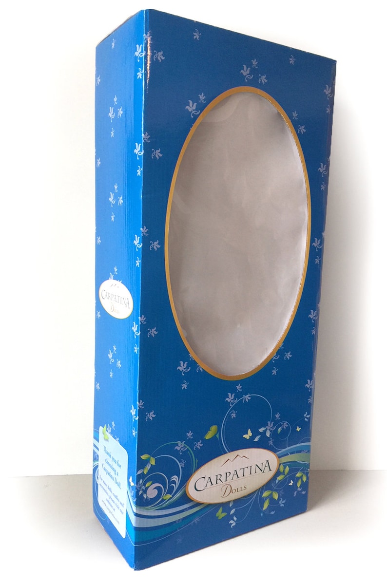 Puede incluir: Una caja rectangular azul con una ventana ovalada, dise&ntilde;ada para mu&ntilde;ecas. La caja presenta un patr&oacute;n floral y las palabras "CARPATINA DOLLS" en un emblema ovalado. La caja tiene un borde dorado alrededor de la ventana ovalada.