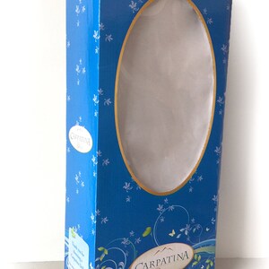 Op de afbeelding: Een blauwe kartonnen doos met een wit ovaal venster dat een pop in de doos laat zien. De doos heeft een wit en blauw bloemenpatroon en de tekst "Carpatina Dolls" onderaan.