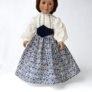 May include: A doll wearing a white blouse with blue embroidery and a navy blue sash, and a white and blue floral print skirt.