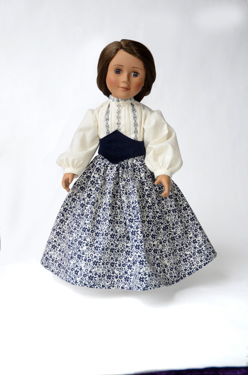 May include: A doll wearing a white blouse with blue embroidery and a navy blue sash, and a white and blue floral print skirt.