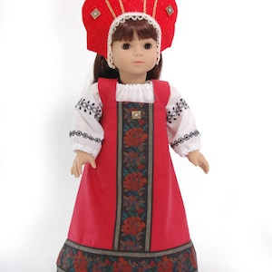 May include: A doll with brown hair wearing a red kokoshnik headpiece with gold trim and square accents. The doll wears a white long-sleeved blouse with black embroidery and a red jumper dress featuring a central panel of dark floral fabric.