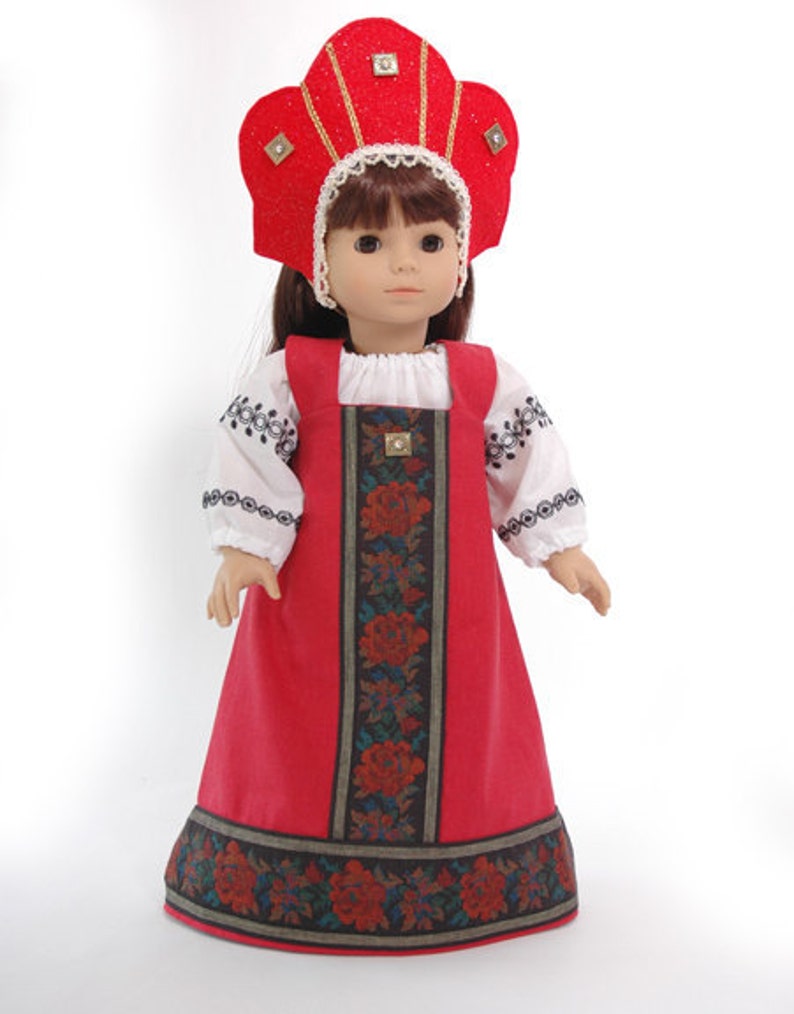 May include: A doll with brown hair wearing a red kokoshnik headpiece with gold trim and square accents. The doll wears a white long-sleeved blouse with black embroidery and a red jumper dress featuring a central panel of dark floral fabric.