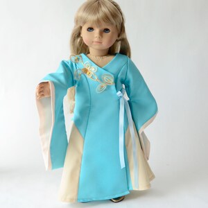 May include: A blue and cream doll dress with gold embroidery and a blue ribbon sash. The dress has long sleeves with cream trim.