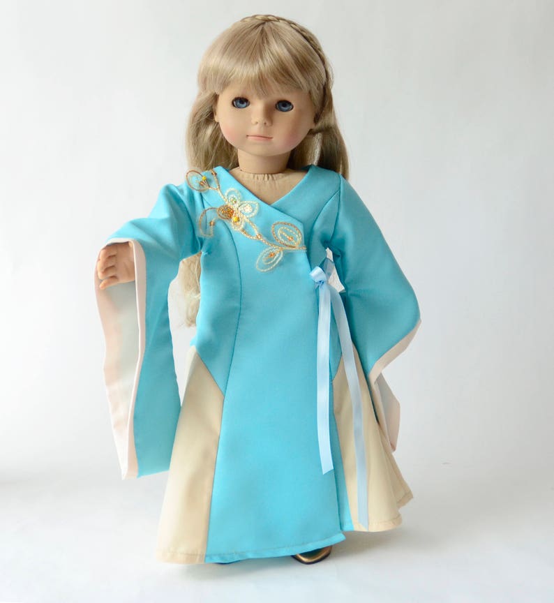 May include: A blue and cream doll dress with gold embroidery and a blue ribbon sash. The dress has long sleeves with cream trim.