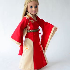 May include: A doll wearing a red and cream dress with gold embroidery and a red sash. The doll has long blonde hair and blue eyes.