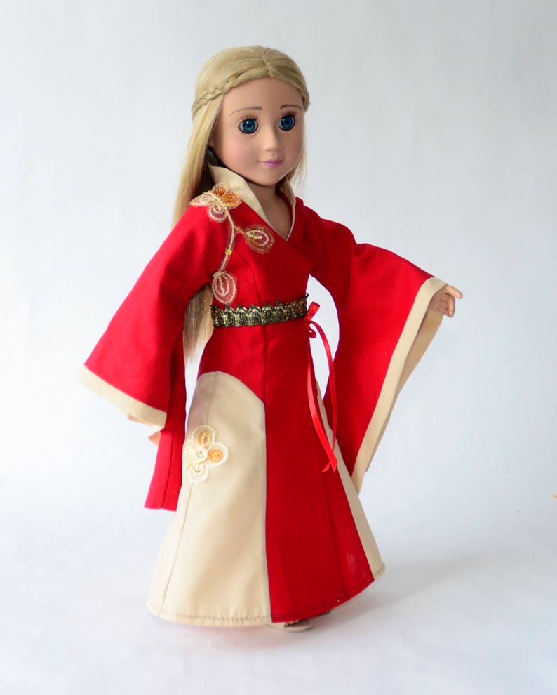 May include: A doll wearing a red and cream dress with gold embroidery and a red sash. The doll has long blonde hair and blue eyes.