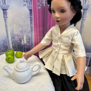 May include: A doll wearing a cream-colored blouse and a black skirt. The doll has black hair and is wearing a silver headband. A white teapot and three green apples sit on a white tablecloth.
