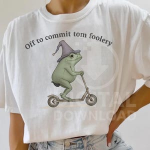 May include: White t-shirt featuring the text "Off to commit tom foolery" and a cartoon frog on a scooter. The frog is wearing a purple wizard's hat. The frog and scooter are in green and grey tones.