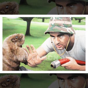 May include: A print featuring a person wearing a camouflage bucket hat and a white t-shirt, holding a red hose with a silver nozzle, gesturing toward a brown furry groundhog standing on a green grassy lawn with trees in the background.