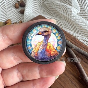 May include: A round, black wooden button featuring a colorful illustration of an ostrich. The ostrich has a tan head, orange beak, and a vibrant display of yellow and orange feathers. The background has a stained glass design with the text "johill design".