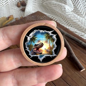 Op de afbeelding: Ronde houten knoop met een dinosaurusscène. De knoop toont een gedetailleerde illustratie van een Tyrannosaurus Rex met open mond, palmbomen en een zonsondergang. De tekst "johill design" staat op de knoop.
