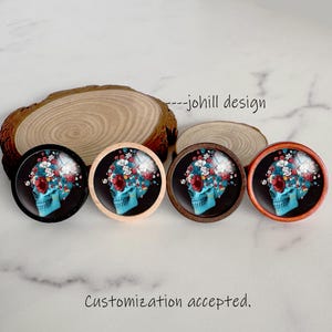May include: Four round decorative objects with a skull design. The skulls are turquoise with floral accents in red, white, and blue. The objects have different colored wooden frames. The text "--johill design" and "Customization accepted" are visible.