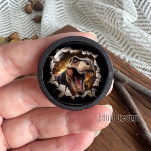 Op de afbeelding: Een zwarte ronde knop met een 3D-illustratie van een Tyrannosaurus Rex die door een gescheurd oppervlak breekt. De dinosaurus heeft bruine en bruine huid, met een open mond. De tekst "johill design" is rechts zichtbaar.