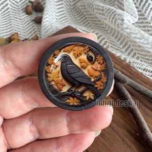 May include: A round, dark wooden carving featuring a detailed raven perched on a branch, surrounded by autumn leaves. The raven is black, and the leaves are shades of orange and brown. The carving has a textured, three-dimensional appearance.