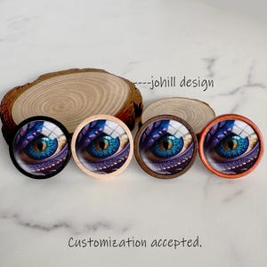 May include: Four round decorative buttons featuring a close-up of a dragon's eye with blue, purple, and gold tones. The buttons have different colored frames: black, natural wood, dark brown, and orange. The text "---johill design" is visible.