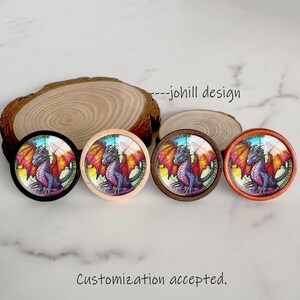 May include: Four round decorative buttons featuring a stained-glass dragon design. The dragon is purple with orange and yellow wings. The buttons have black, light wood, dark wood, and orange frames. Text reads "--johill design" and "Customization accepted."