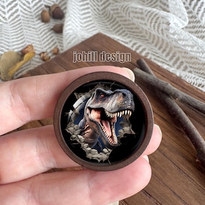 Op de afbeelding: Een ronde houten knoop met een gedetailleerde illustratie van een Tyrannosaurus Rex die door een muur breekt. De dinosaurus heeft scherpe tanden en een dreigende uitdrukking. De tekst "johill design" is op de knoop gedrukt.