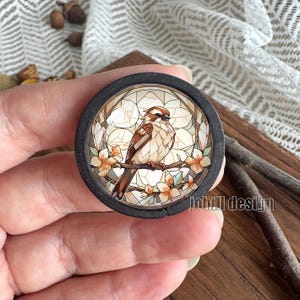 Könnte beinhalten: Ein runder Holz-Ansteckpin mit einem Buntglas-Design, das einen braunen und beigen Vogel auf einem Ast mit pfirsichfarbenen Blüten zeigt. Der Pin hat einen dunkelgrauen Rand und den Text "johill design".