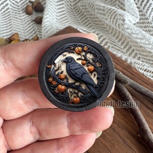 May include: A round, dark gray decorative piece featuring a detailed carving of a black raven perched on a branch with orange pumpkins. The raven is set against a cream-colored background, with the text "johill design" on the right side.