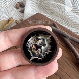 Op de afbeelding: Een ronde houten knoop met een gedetailleerde illustratie van een Tyrannosaurus Rex die door een gescheurd oppervlak breekt. De knoop heeft de tekst "johill design" bovenaan. De dinosaurus wordt afgebeeld met open kaken en scherpe tanden.