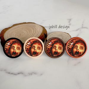 May include: Four round, horror-themed cabinet knobs with a woman's face screaming in terror. The knobs have black, light pink, dark brown, and orange wooden frames. The image is set on a light-colored surface.
