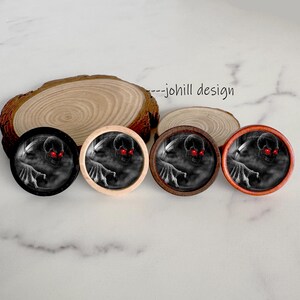 May include: Four round decorative buttons with a spooky skull design. The skulls have glowing red eyes and are set against a dark, misty background. The buttons have black, light wood, dark wood, and orange rims. The text "--johill design" is visible.