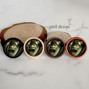 May include: Four round, wooden-framed decorative buttons featuring a scary, green-skinned face with glowing white eyes and sharp teeth. The frames are black, light wood, dark wood, and orange. The text "--johill design" is visible.