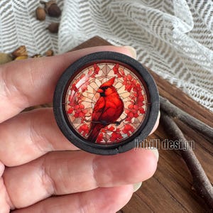 May include: A circular, black wooden item with a stained-glass effect image of a red cardinal bird on a branch with red flowers. The background has a light orange and white pattern. The text "johill design" is visible.