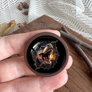 Op de afbeelding: Een ronde, bruine houten knoop met een gedetailleerde illustratie van een Tyrannosaurus Rex die door een gescheurd oppervlak breekt. De tekst "johill design" is op de knoop gedrukt.