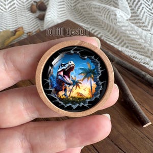 Op de afbeelding: Een ronde houten knoop met een gedetailleerde illustratie van een blauwe Tyrannosaurus Rex in een tropische omgeving. Het ontwerp toont palmbomen, een zonsondergang en de tekst "johill design". De knoop wordt in een hand gehouden.