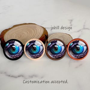 May include: Four round eye-themed decorative objects with blue and green irises, each set in a different colored frame: black, natural wood, dark brown, and orange. The text "--johill design" is visible. The text "Customization accepted." is also visible.