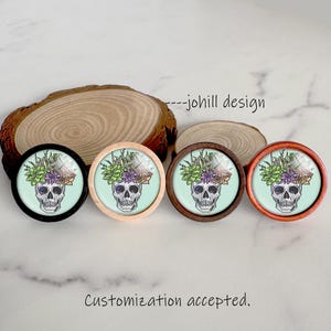 May include: Four round decorative skull designs with floral crowns, each in a different wooden frame: black, natural, dark brown, and orange. The skulls have a light blue background. Text reads "---johill design" and "Customization accepted."