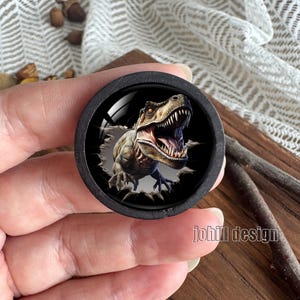 Op de afbeelding: Een ronde, zwarte knoop met een gedetailleerde illustratie van een Tyrannosaurus Rex die uit een donkere achtergrond komt. De dinosaurus is afgebeeld in bruin, beige en grijs, met zijn mond open, en toont scherpe tanden. De tekst "johill design" is zichtbaar.