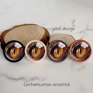 May include: Four round decorative buttons featuring a close-up of a dragon's eye in shades of gold, brown, and orange. The buttons have different colored frames: black, natural wood, dark brown, and orange. The text "---johill design" and "Customization accepted."