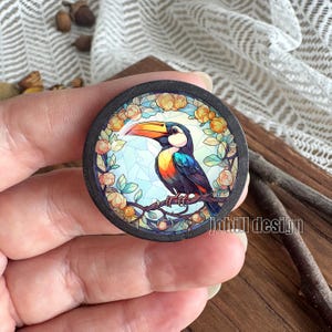 Può includere: Design rotondo di tucano in legno con bordo nero. Il tucano ha un grande becco arancione e giallo, con piume blu, arancioni e nere. Il design è circondato da fiori gialli e foglie verdi. Il testo "johill design" è visibile.