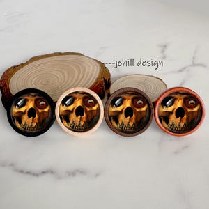 Op de afbeelding: Vier ronde decoratieve knopen met een schedelthema en verschillende houten frames: zwart, licht hout, donker hout en oranje. Elke knoop heeft een gedetailleerde schedelafbeelding onder een helder, gewelfd oppervlak. De tekst "--johill design" is zichtbaar.