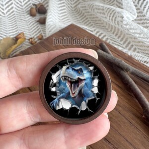 Op de afbeelding: Een ronde houten knoop met een blauwe dinosaurus die door een muur breekt. De dinosaurus heeft een open mond en scherpe tanden. De tekst "johill design" staat op de knoop.