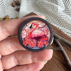 Op de afbeelding: Rond houten ontwerp met een flamingo-ontwerp met glas-in-lood-effect. De flamingo is roze met een zwarte snavel, omringd door roze bloemen en een glas-in-lood-achtergrond. De tekst "johill design" is zichtbaar.