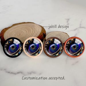 May include: Four round decorative objects with a detailed eye design in blue, gold, and brown. The objects have different colored frames: black, light wood, dark wood, and orange. The text "---johill design" and "Customization accepted" are also visible.