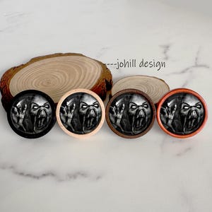 May include: Four round, decorative plugs with a scary monster face design. The plugs have black, natural wood, dark brown, and orange rims. The monster face is black and white with a wide open mouth and outstretched hands. The text "--johill design" is in the background.