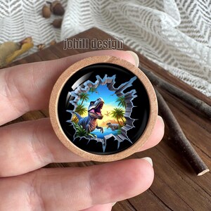 Op de afbeelding: Een ronde houten knoop met een kleurrijke dinosaurusillustratie. De afbeelding toont een blauwe dinosaurus, palmbomen en een blauwe lucht. De tekst "johill design" is ook aanwezig.