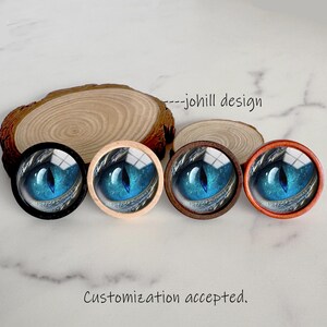 May include: Four round decorative objects with a blue eye design, each with a different colored frame: black, light wood, dark wood, and orange. The text "---johill design" and "Customization accepted" are also visible.
