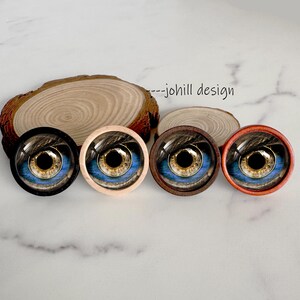 May include: Four round eye cabochons with detailed iris and pupil designs. The eyes have a blue and gold color scheme, with black, light brown, dark brown, and orange frames. The text "--johill design" is visible.