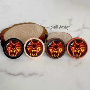 May include: Four round wooden plugs with a fiery demon face design. The plugs have black, light wood, dark wood, and orange wood frames. The image includes the text "--johill design".