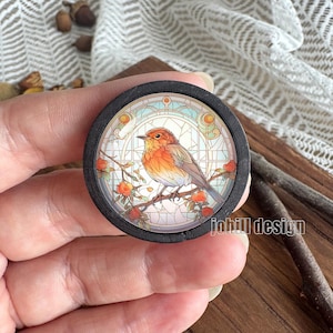 Op de afbeelding: Ronde, zwarte houten pin met een glas-in-lood illustratie van een vogel op een tak met rode bessen. De vogel heeft oranje en bruine veren. De tekst "johill design" is rechtsonder zichtbaar.