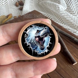 Op de afbeelding: Een ronde houten knoop met een gedetailleerde illustratie van een Tyrannosaurus Rex die door een gescheurd oppervlak breekt. De dinosaurus is grijs en wit, met een open mond en scherpe tanden. De tekst "johill design" is zichtbaar.